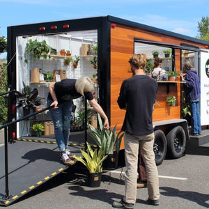 A greenhouse on wheels, filled with houseplants and handmade pottery, being visited by happy shoppers on a sunny day.